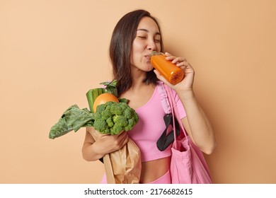 Diet and superfood concept. Healthy dark haired Asian woman drinks homemade orange juice detox drink carries fresh vegetables in paper bag has proper nutrition isolated over brown background