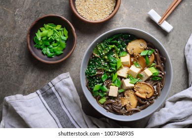 Diet vegetarian bowl of soba noodle soup rich with various sources of vegan protein such as mushrooms, tofu and kale; the meal seasoned with sesame seeds and chives. View from above arrangement.