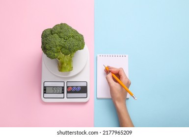 Diet, weight loss, calorie counting concept. Broccoli on a kitchen scale, a hand writes information in a notebook on a blue-pink pastel background. Top view - Shutterstock ID 2177965209