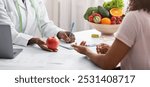 Dietician african woman giving young patient healthy diet tips, holding fresh red apple, clinic interior