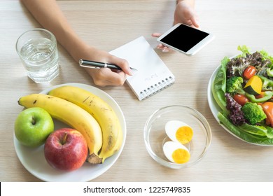 Dieting And Calories Control For Wellness. Woman Using Smartphone Calculate Calories Of Food In Breakfast During Dieting For Lose Weight Program And Take Notes.