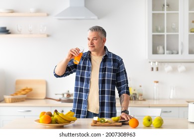 Dieting and nutrition for health. Sporty mature man drinking fresh juice, standing near table full of various fruits in kitchen interior, empty space - Powered by Shutterstock