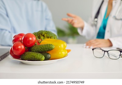 Dietitian Or Nutritionist Giving Consultation To Patient. Close Up Of Plate Of Fresh Raw Vegetables On White Table, With Doctor And Client Talking In Background. Healthy Food, Eating Healthy Concept