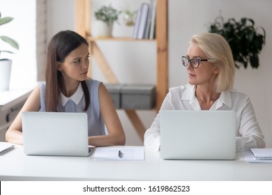 Different Ages Women Colleagues 50s And Young Female Workmates Sit At Office Desk Looking With Enmity At Each Other. Negative Attitude, Conflict At Workplace, Misunderstanding Generational Gap Concept