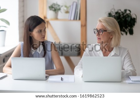 Different ages women colleagues 50s and young female workmates sit at office desk looking with enmity at each other. Negative attitude, conflict at workplace, misunderstanding generational gap concept