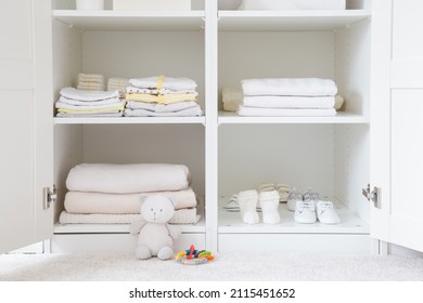 Different baby clothes, shoes and bedclothes on shelves in white opened wardrobe at nursery room. Front view.