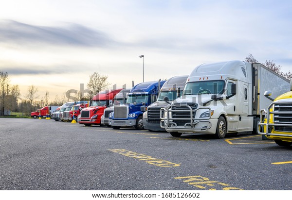 Different big rigs semi trucks with semi\
trailers standing in row on truck stop parking lot with reserved\
spots for truck driver rest and compliance with established truck\
driving regulations