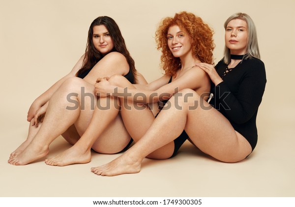 Different Body Types. Diversity Models
Portrait. Blonde, Brunette And Redhead In Black Bodysuits Sitting
On Floor. Group Of Women Posing On Beige Background.
