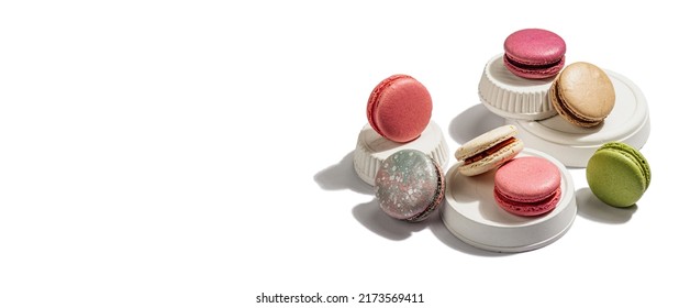 Different colorful French macarons isolated on white background. Sandwich cookies on trendy stands. Assorted confectionery range, hard light, dark shadow, banner format