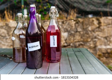 Different coloured glass bottles filled with homemade liquor, with the word 'Mirtillo' (translation: Blueberry) handwritten on sticky labels, standing on garden table