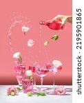 Different elegant champagne glasses with sparkling wine, women hand is pouring rose wine from bottle to coupe glass. Splash, splatter, fly flowers with water drops on the pink background