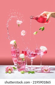 Different elegant champagne glasses with sparkling wine, women hand is pouring rose wine from bottle to coupe glass. Splash, splatter, fly flowers with water drops on the pink background