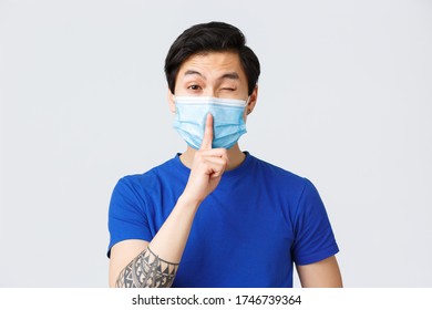 Different Emotions, Lifestyle And Leisure During Coronavirus, Covid-19 Concept. Cheeky Handsome Asian Man In Medical Mask Promise Keep Secret, Shush And Wink Coquettish, Be Silent