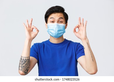 Different Emotions, Lifestyle And Leisure During Coronavirus, Covid-19 Concept. Asian Smiling Guy In Medical Mask Tell Nothing Worry About, Relax, Show Okay Sign And Guarantee Quality, Approve Smth