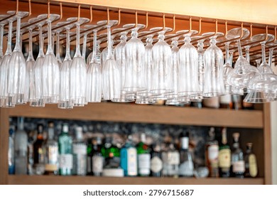 Different empty glasses hang over the bar counter with shelves filled with various bottles of alcohol behind. Alcoholic drink establishment or beverage bar in hotel