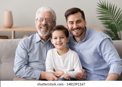 Different generation age caucasian men together at home, smiling grandpa grown up son and small cute grandson sitting on couch in living room looking at camera, multi generational male family concept