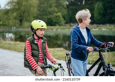 Different Generation Family Communication Concept. Smiling Senior Woman With Her Grandson Walking On A Country Lane With Bicycles With Lake On Background.