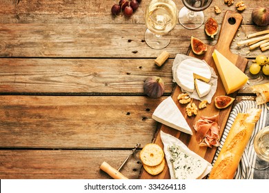 Different Kinds Of Cheeses, Wine, Baguettes, Fruits And Snacks On Rustic Wooden Table From Above. French Tasting Party Or Feast Scenery. Layout With Free Text Space. 