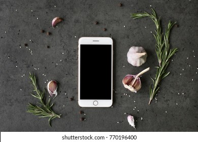 Different Kinds Of Spices: Rosemary, Salt, Pepper And Garlic On The Dark Slate, Stone Or Metal Background With Phone Mockup With Empty Screen. Food Flatlay Top View With Copy Space For Text