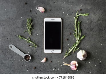 Different Kinds Of Spices: Rosemary, Salt, Pepper And Garlic On The Dark Slate, Stone Or Metal Background With Phone Mockup With Empty Screen. Food Flatlay Top View With Copy Space For Text