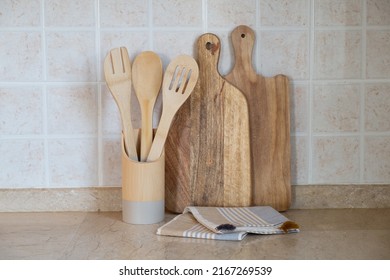 Different Kitchen Utensil Made Of Wood In A Holder, Cutting Board And A Blanket On Marble Tabletop. Copy Space For Text, Close Up, Wall With Beige Facing Tile Background.