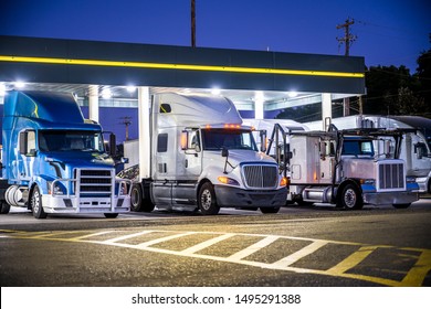 Different make and models big rigs semi trucks with semi trailers standing on the truck stop parking lot under the lighted shelter in night and comply with the movement according to the schedule