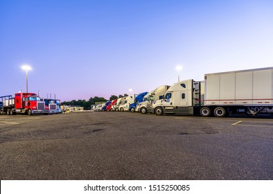 Different Make And Models Big Rigs Semi Trucks With Semi Trailers Standing In Row On Truck Stop Parking Lot For Rest And Comply With The Movement According To The Schedule For Successful Delivery