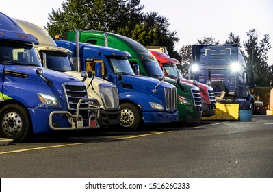 Different Make And Models Big Rigs Semi Trucks With Semi Trailers Standing In Row On Truck Stop Parking Lot For Rest And Comply With The Movement According To The Schedule For Successful Delivery