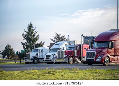 Different models of big rigs semi trucks tractors with car hauler and refrigerator and dry van semi trailers standing in row on the truck stop industrial parking lot for the rest at twilight time