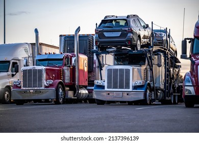 Different models of big rigs semi trucks tractors with car hauler and refrigerator and dry van semi trailers standing in row on the truck stop industrial parking lot for the rest at twilight time