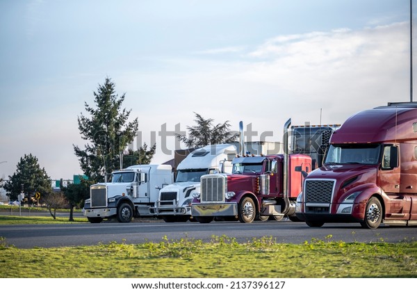 Different models of big rigs semi trucks tractors\
with car hauler and refrigerator and dry van semi trailers standing\
in row on the truck stop industrial parking lot for the rest at\
twilight time