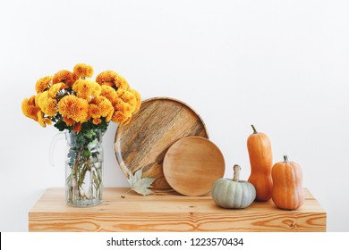 Different Multicolored Decorative Pumpkins, Wooden Plates Or Dishes And Vase With Yellow Chrysanthemums Flowers On A Wooden Table On A Background Of White Wall. Autumn Or Fall Home Interior Decor.