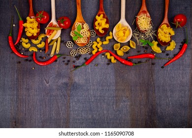 Different pasta in spoons on a dark wooden background, top view. Raw pasta, chili, tomatoes and basil close-up.