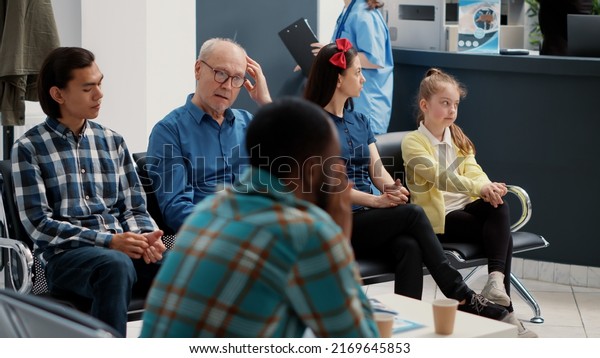 Different\
people waiting in row at hospital reception lobby to be called in\
office at checkup visit examination with specialist. Waiting room\
area for medical consultation at busy\
clinic.
