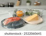 Different products with plastic food wrap on white marble table in kitchen