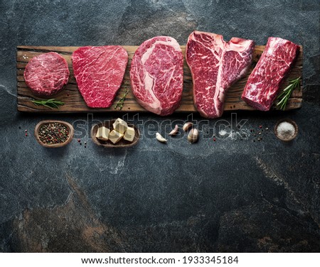 Different raw beef steaks on the wooden board on the grey table. Top view.