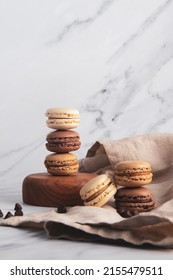 Different shades of brown macaroons piles on top of each other with marble background and cream napkin with chocolate drops scattered around. 
