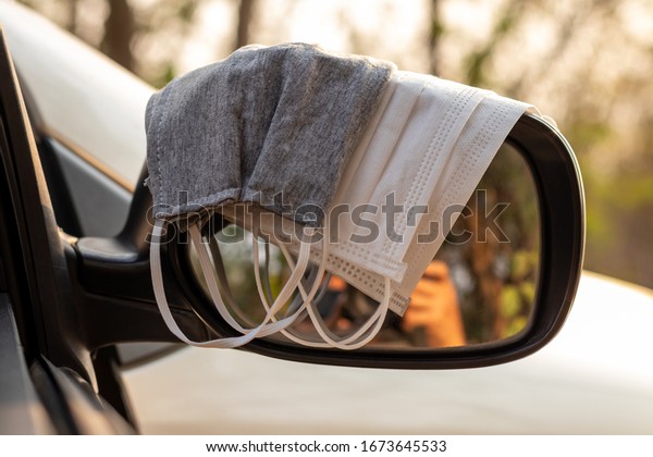 Different types of hygienic masks, both white\
and gray, are placed on the side mirrors of the car, with the\
countryside blurred in the\
background.