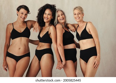 Different Women Of All Ages Celebrating Their Natural And Aging Bodies. Four Confident Women Smiling Cheerfully While Wearing Black Underwear And Standing Together Against A Studio Background.