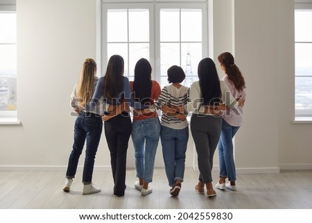 Different women in casual clothes pose with their backs to camera hugging each other like best friends. Women who show their support and equality stand in row in bright room and look out the window.