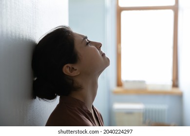 Difficult life moment. Tired stressed indian woman sit leaning against white wall in light room try to cope with problems. Side profile view of young hindu lady face with closed eyes think breath deep