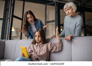 Difficult Relationship. Daughter, Carried Away By A Smartphone, Looking At The Screen, Stopping The Conversation Of Her Grandmother And Mother, Who Are Upset.