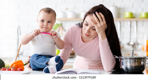 Difficulties Of Maternity And Freelance. Cute Baby Biting Pen, Tired Mom Sitting Aside, Kitchen Interior, Panorama