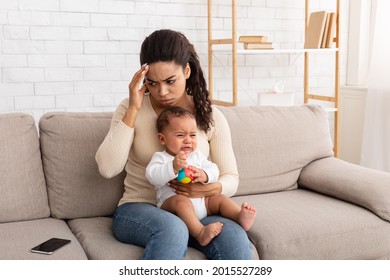 Difficulties Of Motherhood. African American Mother With Crying Infant Baby Suffering From Headache Pain Sitting On Couch At Home. Ill Mom Touching Aching Head Caring For Little Son Indoor