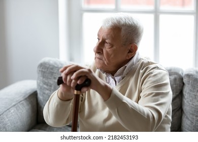 Difficulties in walking. Sad elderly male sitting on sofa feeling pain in legs as result of chronic age related diseases. Pensive older man holding wooden cane give rest to feet tired after long walk