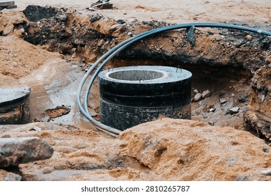 digging at a construction site where a large circular concrete structure is visible alongside exposed pipes. The ground is uneven with dirt and sand scattered around the area.
