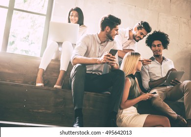 Digital age students. Group of cheerful young people communicting while holding different gadgets and sitting close to each other on steps 