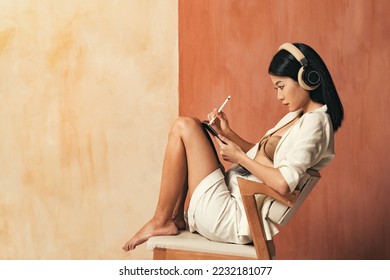 Digital artist and art. Side view of concentrated asian woman graphic designer in headphones using tablet, drawing illustrations with stylus working online sitting on chair on copy space background - Powered by Shutterstock