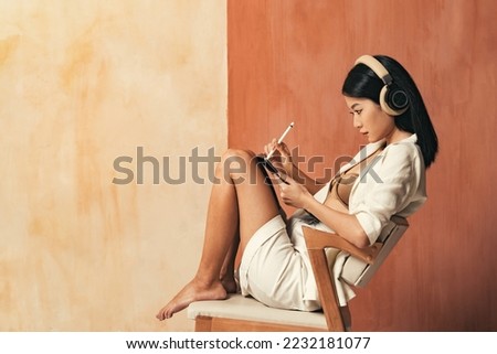 Digital artist and art. Side view of concentrated asian woman graphic designer in headphones using tablet, drawing illustrations with stylus working online sitting on chair on copy space background