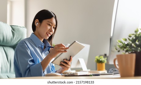 Digital Artist. Smiling Asian Female Graphic Designer In Earbuds Using Tablet, Drawing With Stylus Working Remotely Online Sitting On Chair At Desk In Modern Office. Profile Side View Banner Panorama